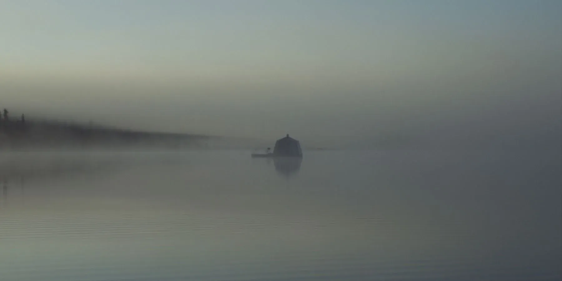 A boat on still water in heavy mist, a treeline faint behind it