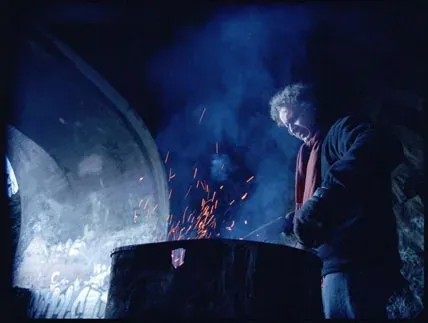 A man tending a fire in a metal drum at night, sparks rising